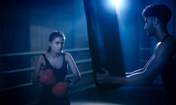 Indian young adult gen z angry fit boxer woman lady wear gloves do hand fist boxing punch beat on bag indoor dark room place modern male sports trainer female girl learn self defense martial art skill - Powered by Adobe