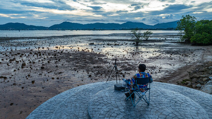 A man sits alone eating in a folding chair on a stone platform, overlooking the rocky intertidal flats, camera at his side, and the peaceful sea in the distance. A quiet moment of solitude at sunset.