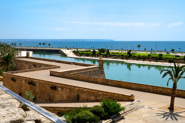 Historic stone fortress walls with turquoise water canal and palm trees overlooking the Mediterranean Sea in Palma de Mallorca, scenic coastal view blending heritage architecture and seaside beauty un