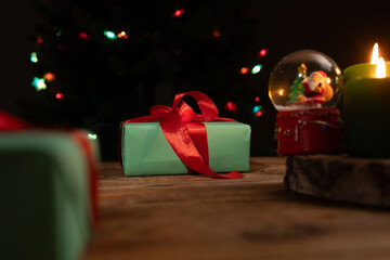 Wrapped Christmas presents sit on a rustic wood surface, alongside a Santa snow globe and lit candles. Christmas tree lights shine in the dark background, creating a festive mood.