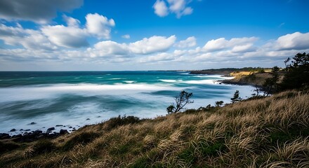 Scenic Coastal Landscape with Deep Blue Ocean, Crashing Waves, and Grassy Foreground Under Partly Cloudy Sky