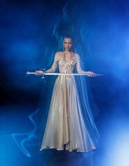 real person sexy face fantasy woman queen holding magic glowing sword in hands. Black dark night room blue moon light fog. Gothic girl warrior with sharp blade shiny dress mixed light art photography