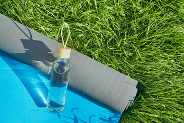 Reusable glass bottle of water and yoga mat on green lawn