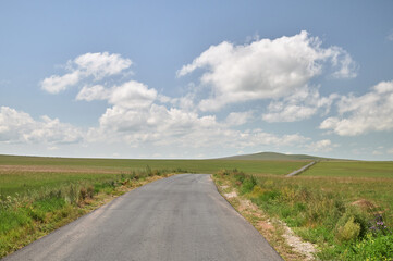 A winding gravel road leads through a vast green landscape under a bright blue sky with scattered white clouds