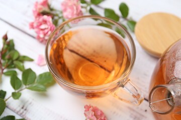 Tasty rose tea in glass cup, flowers and leaves on white wooden table, closeup