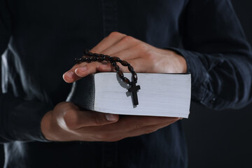 Priest with rosary and Holy Bible on black background, closeup