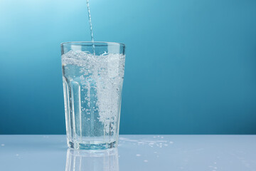 Pouring refreshing soda water into glass at white mirror table against light blue gradient...