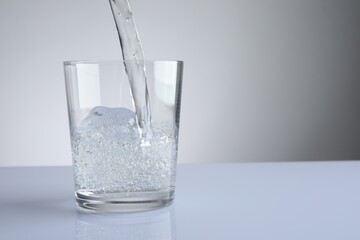 Pouring refreshing soda water into glass at white mirror table against light grey gradient...