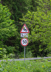A vertical shot of road signs against a backdrop of a green forest. The signs warn of a series of dangerous turns and a speed limit of 50 km/h.