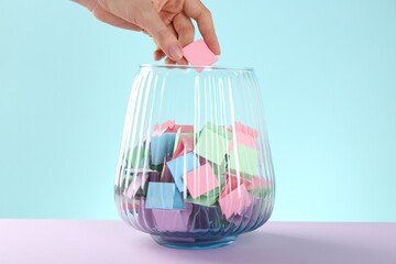 Woman taking pink paper piece from glass jar on color background, closeup