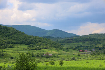 Vast green hills covered with fields and trees stretch towards the horizon. In the valley, scattered rural houses are visible, with gray clouds floating above them, creating an atmosphere of peace and