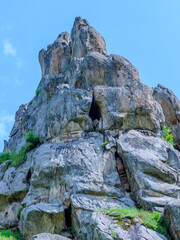 An impressive rock formation of massive stone structures rises towards a blue sky. Green vegetation sprouts on its slopes, adding contrast to the gray surface of the stones.