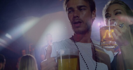 Man wearing white tee, necklace raising beer mug showing thumbs-up in bar with colored stage lights