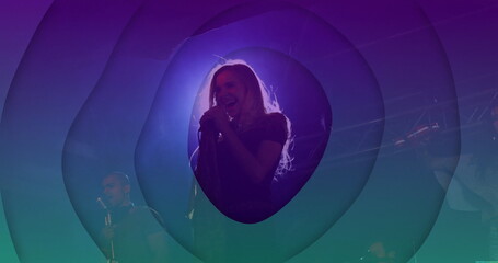 Singing blonde female lead singer holding handheld microphone on stage, with colored lights haze