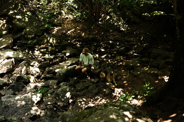 A man sits on rocks with his German Shepherd in a shaded forest, surrounded by mossy stones and dappled sunlight. Stara planina, Serbia