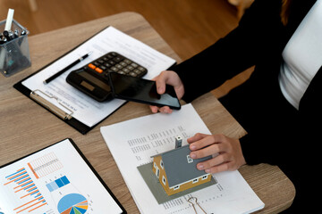 Adult Asian hands holding house model smartphone calculator documents wooden desk real estate financial planning mortgage investment property analysis home buying selling