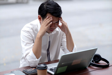 Asian adult businessman feeling overwhelmed with work using laptop computer outdoors experiencing burnout or financial problem