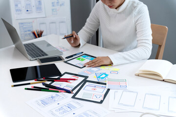 Asian adult woman UX UI designer prototyping mobile app interfaces using tablet laptop creating user experience at modern office desk