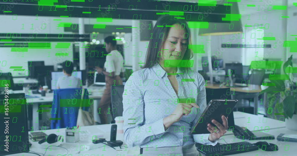 Aufkleber holding tablet and tapping screen at open-plan office desk, with monitors and binary code overlay