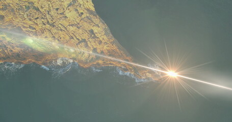 Rocky peninsula extending into calm teal ocean at sunset from above, with sunbeam lens flare
