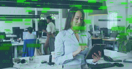 Holding tablet and tapping screen at open-plan office desk, with monitors and binary code overlay