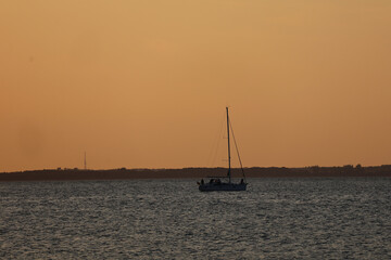 Sailboat with retracted sail drifting on sea near beach at sunset, maritime landscape