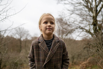 Pensive child wearing a coat stands amidst serene forest surroundings during winter, embodying...