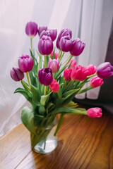 Vase with fresh pink and purple tulips on a wooden windowsill. Delicate spring flowers and soft daylight create a romantic, cozy, and cheerful atmosphere. Ideal for decor and seasonal concepts