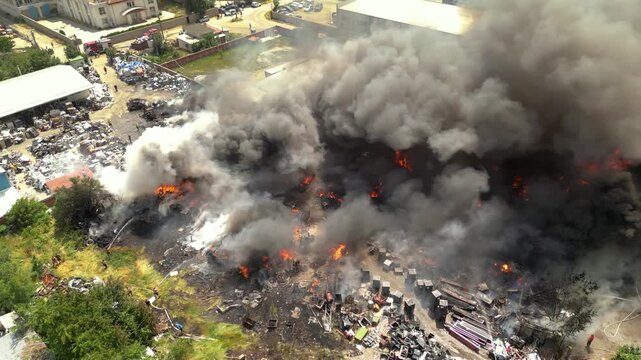 Environmental disaster scene of recycling plant fire. Aerial drone shot shows thick smoke, toxic fumes, burning garbage and firefighters fighting pollution crisis.
