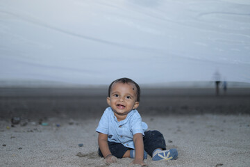 Adorable baby boy sitting on sandy beach next to decorative cubes with balloons spelling “ONE”...