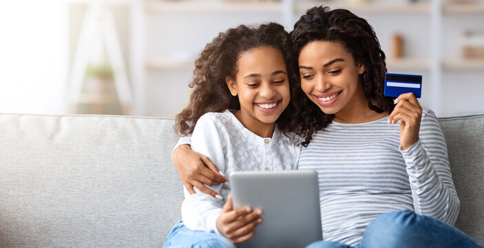 Cute african american mother and kid shopping together on digital tablet, holding credit card, sitting and hugging on couch at home, looking at pad screen and smiling, order food, copy space