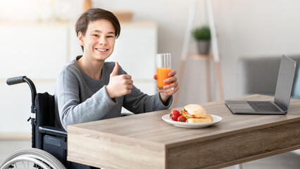 Happy disabled teenager having breakfast, holding glass of juice, showing thumb up gesture at home....
