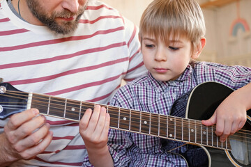 Handsome dad teaches his cute son to play the guitar sitting on sofa in the living room at home. Happy Father's Day. Father teaching his child to play guitar. Kid Learning music skill. Art education