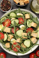 Tasty salad with croutons, cucumbers, tomatoes and arugula on black table, flat lay
