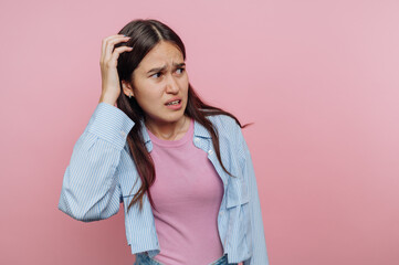 Confused young woman with long hair stands against a pink background while scratching her head in...