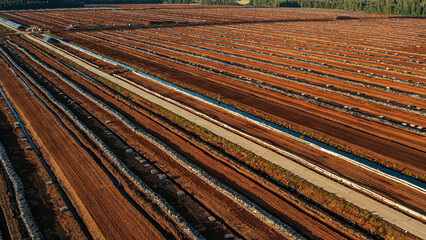 Aerial view of a large peat bog harvesting site with long parallel rows of extracted peat, wooden pallets, and covered stacks stretching across the landscape.