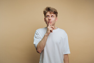 Young man in white t-shirt holding finger to lips with neutral background, conveying a sense of...