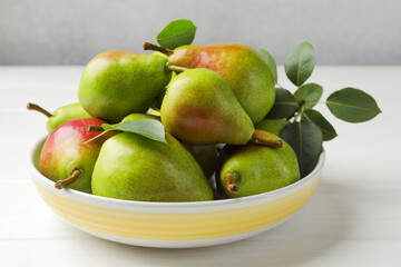 Fresh ripe pears in bowl and green leaves on white wooden table, closeup