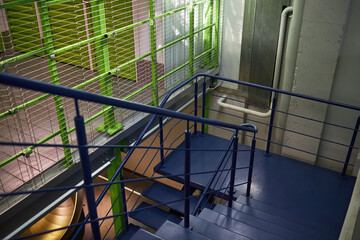 Empty modern staircase with blue metal railings and green mesh fencing leading down to lower level in contemporary building, industrial pipes and concrete walls visible in background