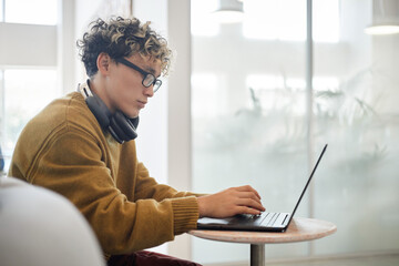 Young asian man with curly hair and glasses sitting at table using laptop, wearing headphones around neck, focusing on typing, modern office environment visible in background