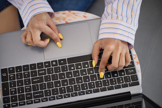 Young adult woman typing on laptop keyboard, hands with yellow painted nails pressing keys, striped shirt sleeves visible, working or studying on portable computer