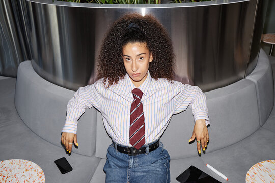 Portrait of young adult woman sitting on modern sofa, looking directly at camera with serious expression, resting arms on backrest, two smartphones and marker nearby - Powered by Adobe