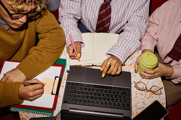 Three young adults collaborating at table, multiethnic group working on laptop and writing in notebooks, one holding reusable coffee cup, hands and torsos visible, business teamwork concept