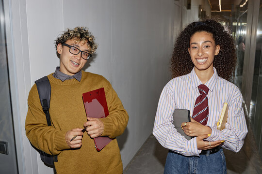Two young adults, Asian man with curly hair and biracial woman with long curly hair, standing in hallway holding folders and digital tablet, smiling at camera - Powered by Adobe