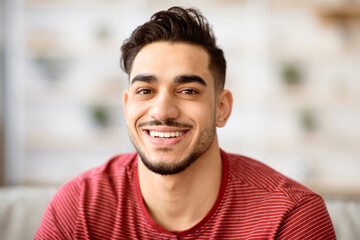 Fototapeta premium Closeup portrait of cheerful handsome arab guy smiling at camera while chilling at home, copy space. Positive young middle-eastern bearded man posing at home, wearing casual outfit