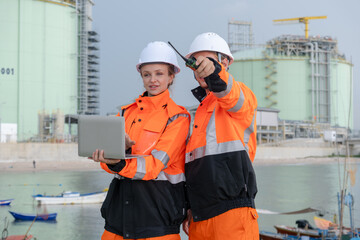 Workers coordinate operations near industrial facility while using laptop and radio at a waterfront...