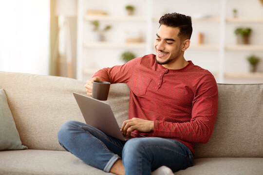 Smiling arab guy chatting with ladies online and drinking coffee, using dating website on laptop while resting on sofa at home. Happy muslim man watching movies or funny videos on notebook, copy space