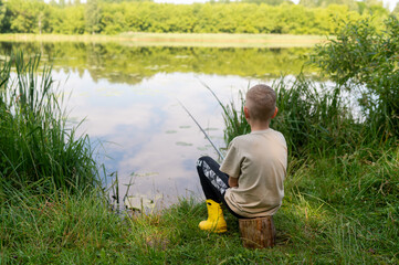 Blond boy sitting on the lake shore fishing with a rod on a sunny summer day