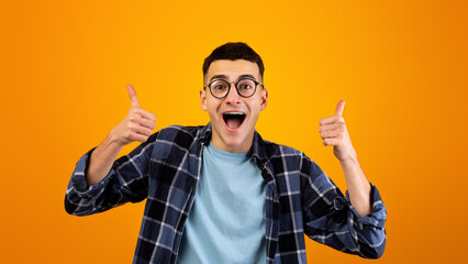 Funky young guy in glasses showing thumbs up with both hands, recommending something on orange...
