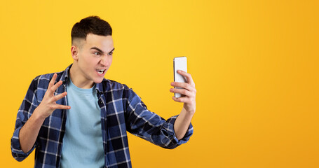 Terrible connection. Angry millennial man looking at his smartphone in rage over orange studio...